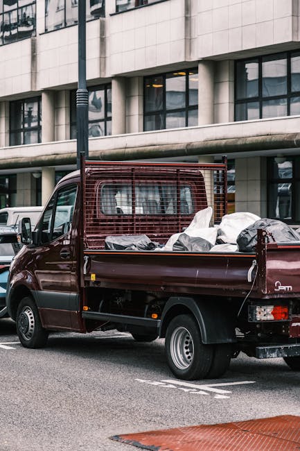 A maroon flatbed utility truck parked on an urban street in front of a modern building with large glass windows and concrete panels. The truck's cargo bed is loaded with various discarded items includ