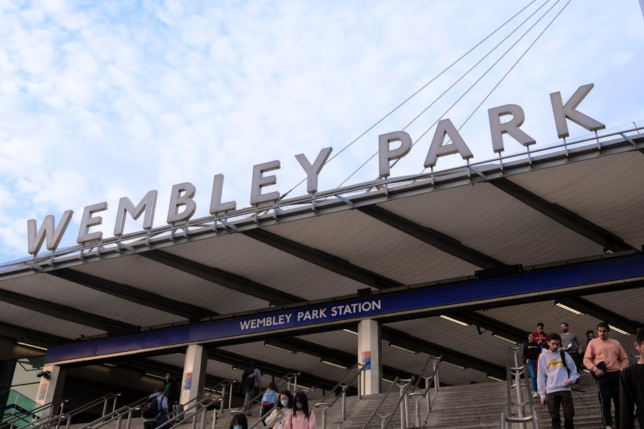 An upward view of Wembley Park Station entrance, with large, metallic, three-dimensional letters spelling 'WEMBLEY PARK' mounted on the station's canopy roof. The structure features a modern, flat roo